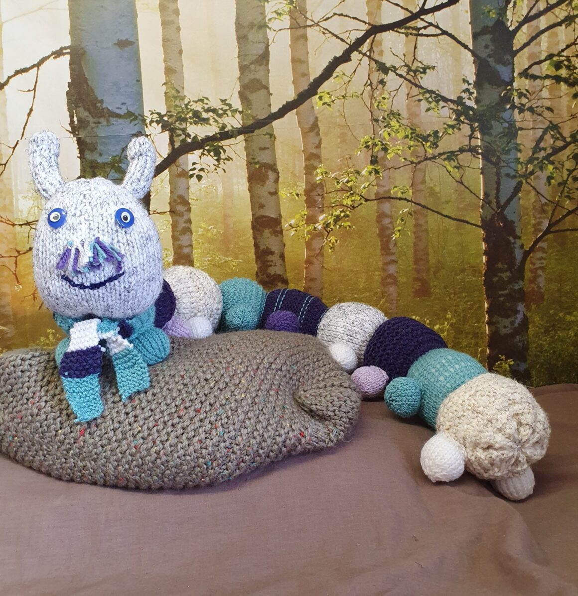 Knitted caterpillar sat on a cushion in front of a woodland image