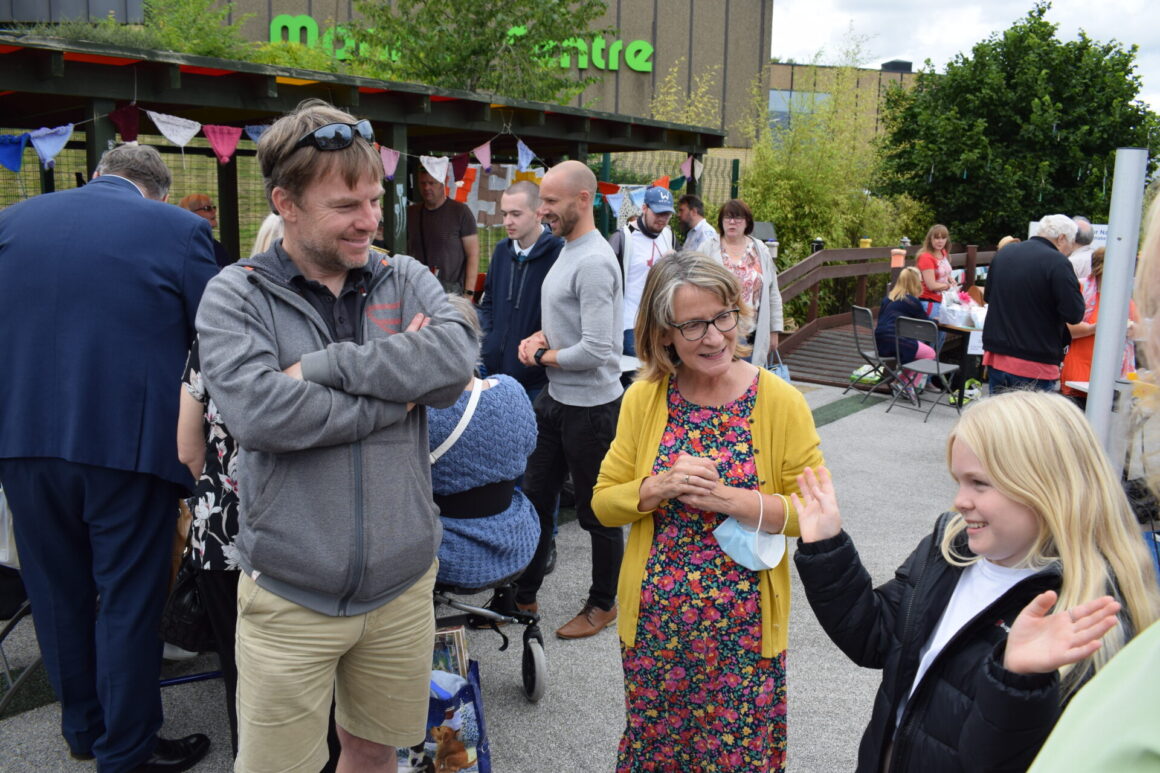 Groups of people standing. Young girl has her arms spread out showing her 'sign name' for BSL