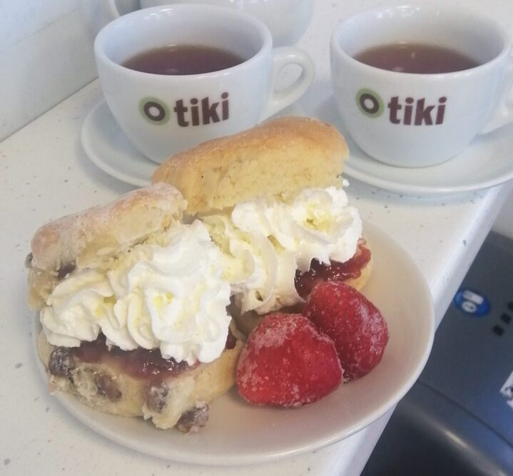 Two cream scones on a plate with two cups of tea behind