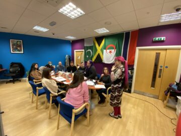 Meeting room with people seated around table. There are flags on the wall. Hannah stands at the front leading the training