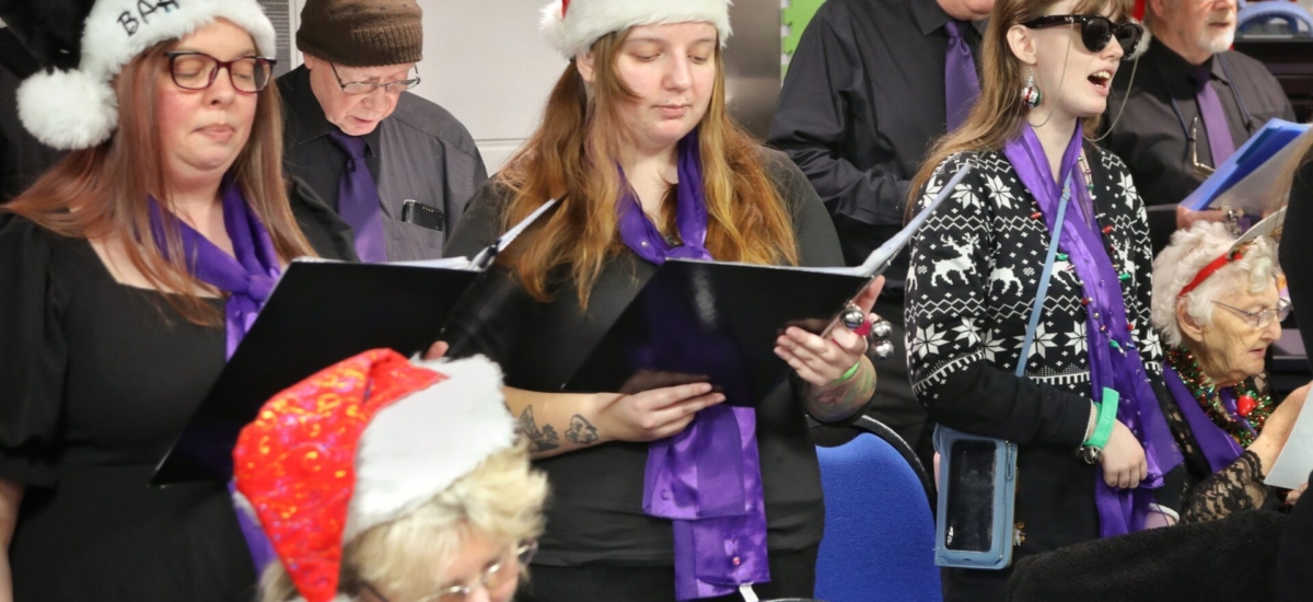 Photo shows the Sensory Singers Choir wearing black clothes and purple ties and scarves. Most are wearing Santa hats. They are holding songbooks and singing. 