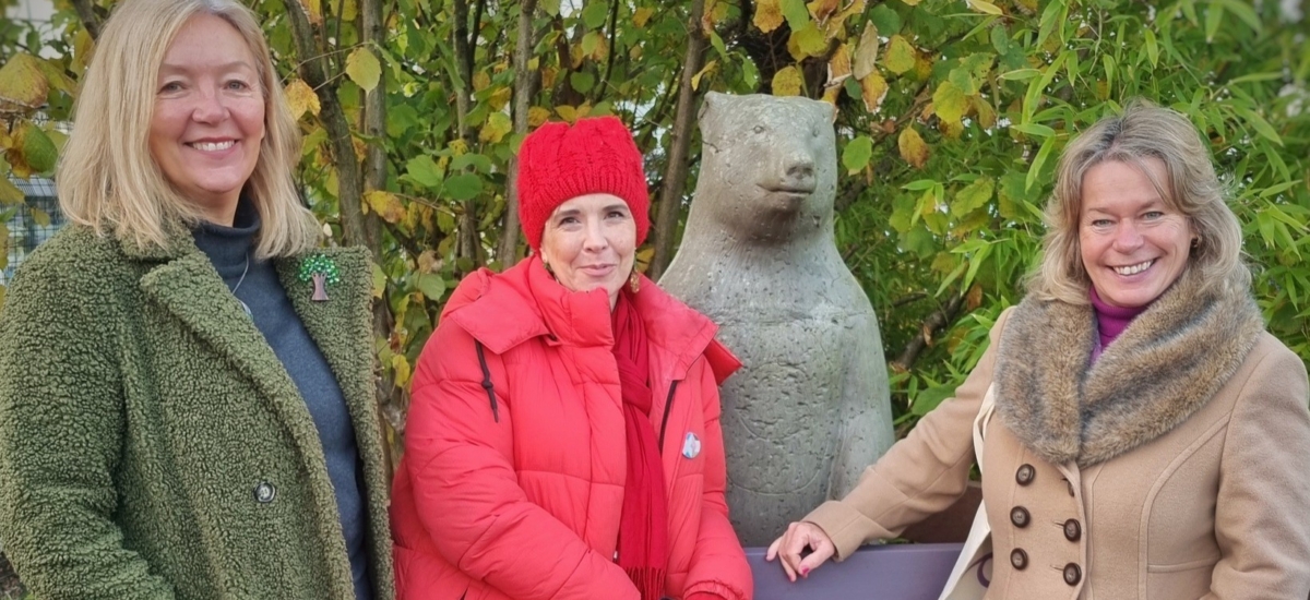 Photo show three people standing beside the bear sculpture in our Sensory Garden. They are wrapped up in hats and coats. The bear is equal height with them all. 